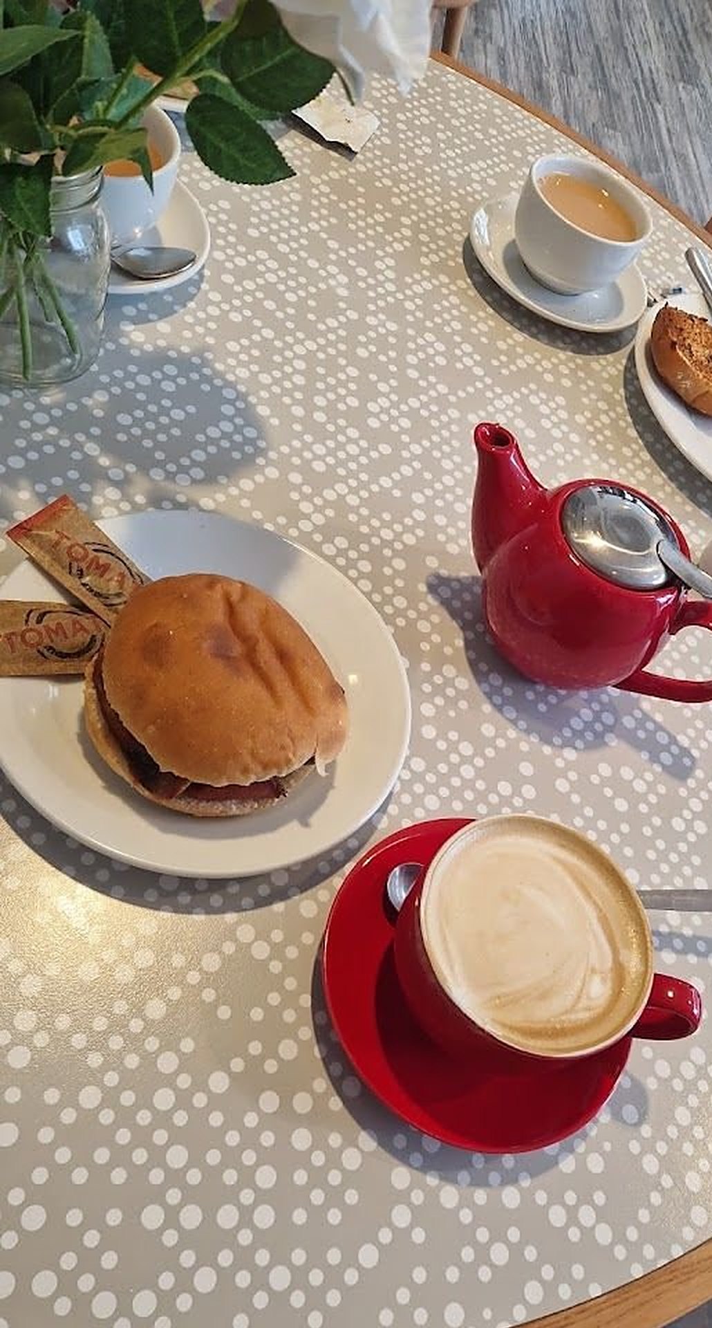 A red teapot, a cappuccino in a red cup, a toasted bun on a white plate, on a grey spotted round table