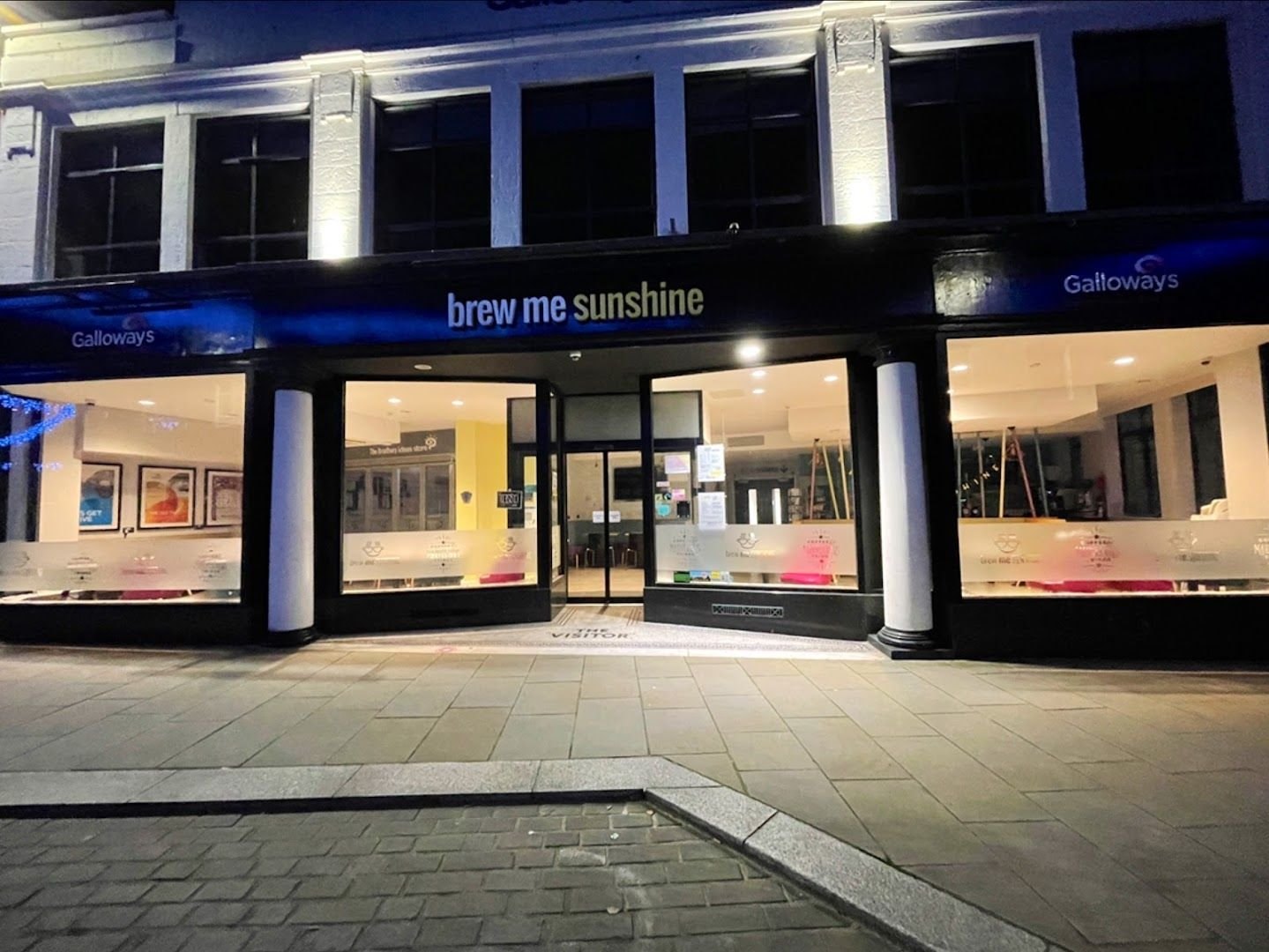 Brew Me Sunshine at night: a wide lit glass shopfront under a dark fascia reading ‘brew me sunshine’ with Galloways signs either side