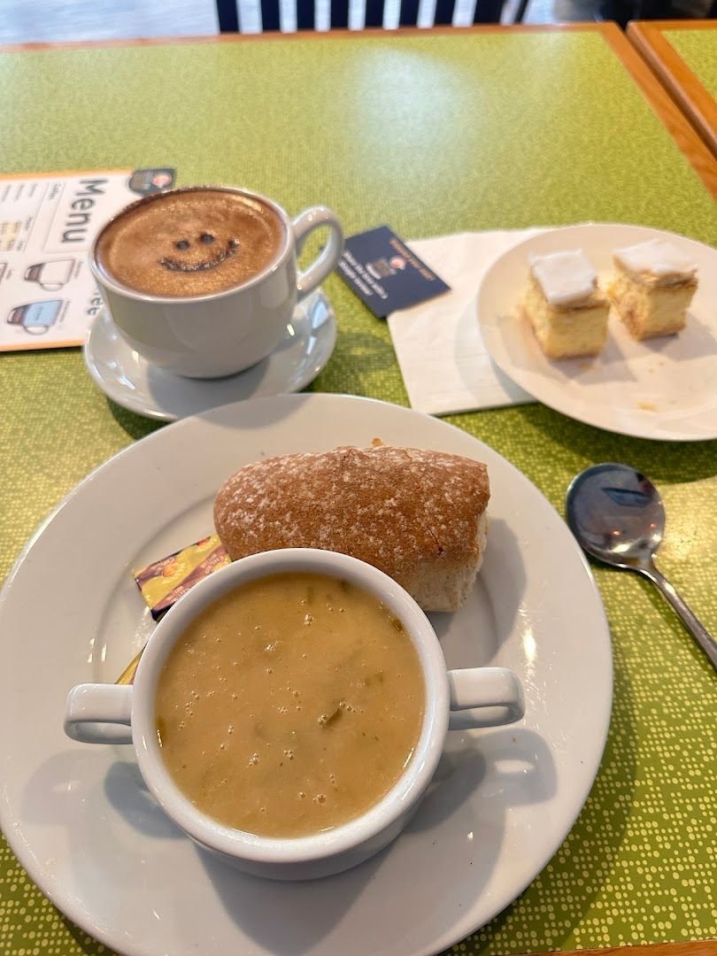 A coffee with a smiley face dusted on the foam, a bowl of soup with a bread roll, and two iced sponge squares on a green table